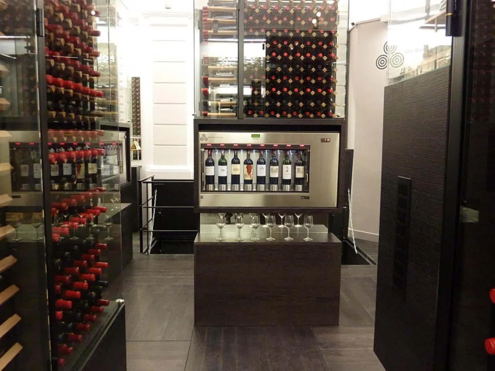 Contemporary interior of Max Bordeaux Wine Gallery & Cellar featuring red wine displays, a central tasting counter with wine glasses, and illuminated bottle presentations.