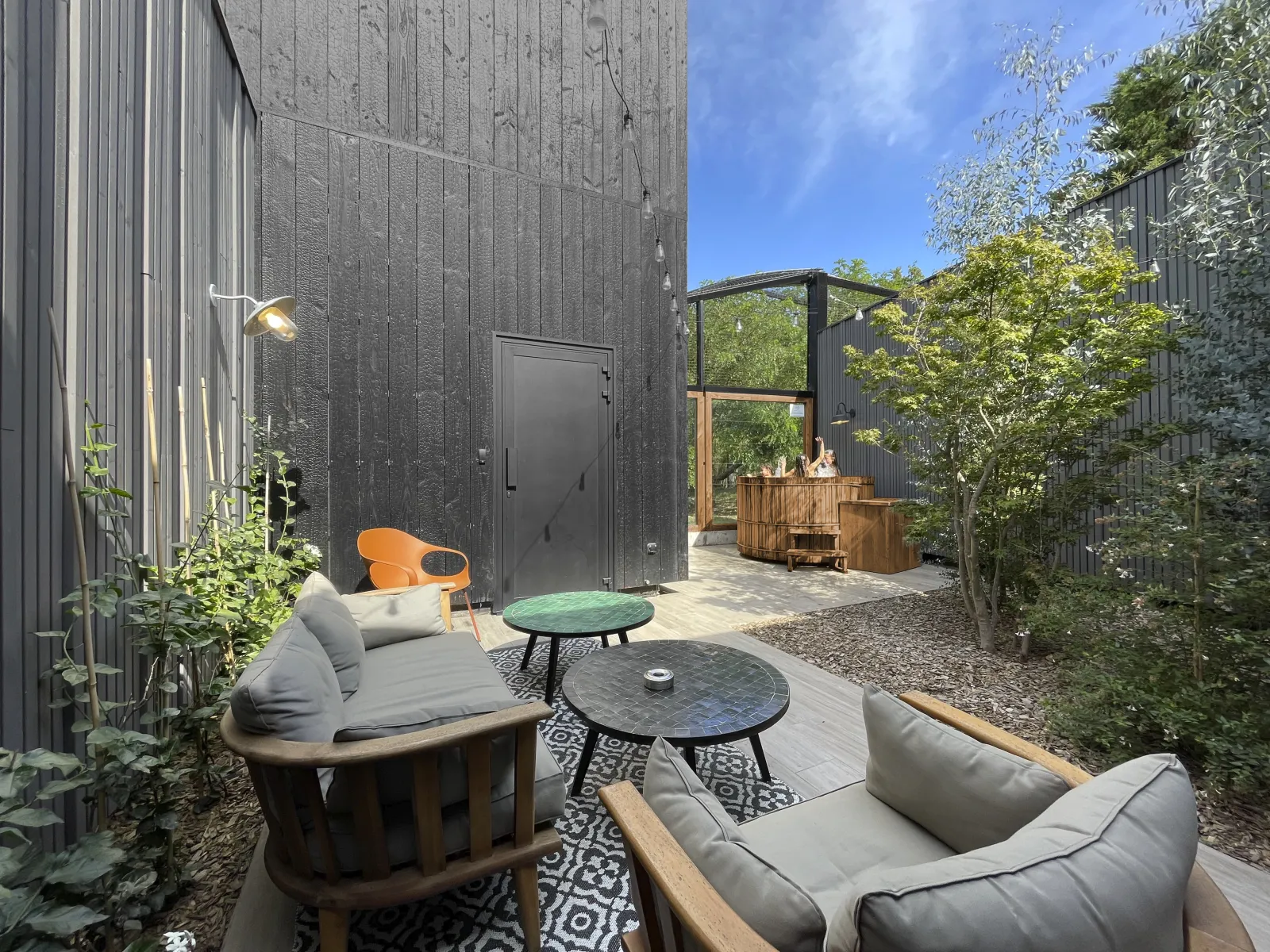 Modern outdoor terrace in Bordeaux featuring designer furniture, black metal and wooden walls, integrated greenery, low tables and comfortable seating under blue sky.