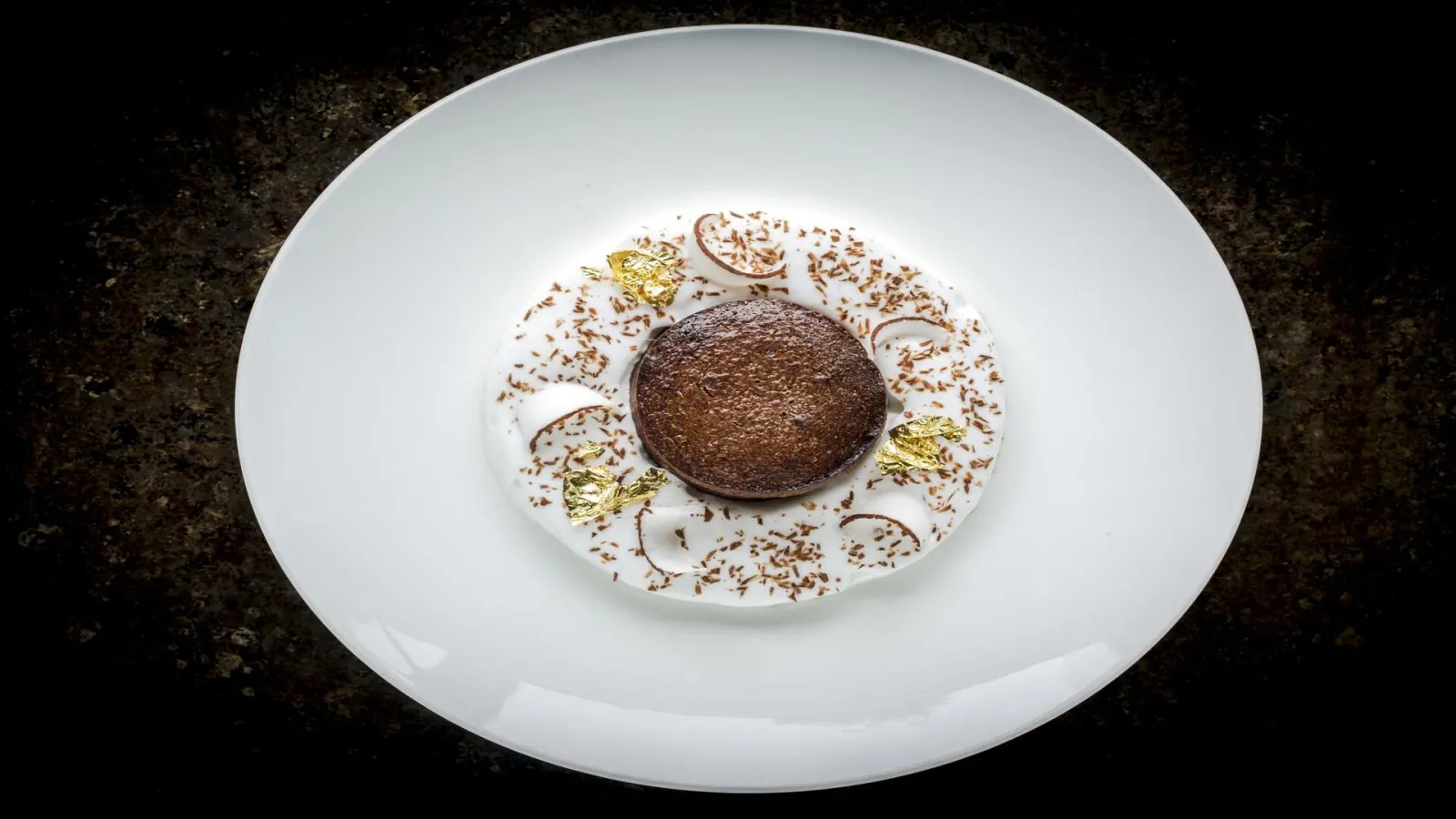 White plate featuring a gourmet dessert with a centered chocolate mousse surrounded by cocoa powder and gold leaf, from Le Bordeaux restaurant