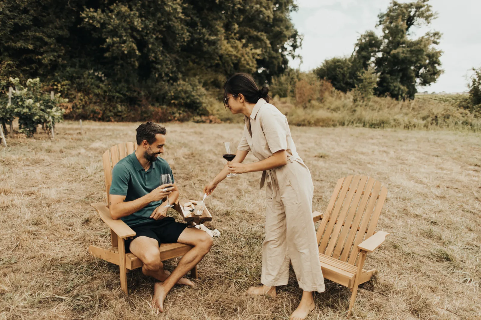 Couple enjoying red wine sitting in wooden chairs in the Saint-Emilion vineyards near Bordeaux, with trees and vineyard rows in the background.