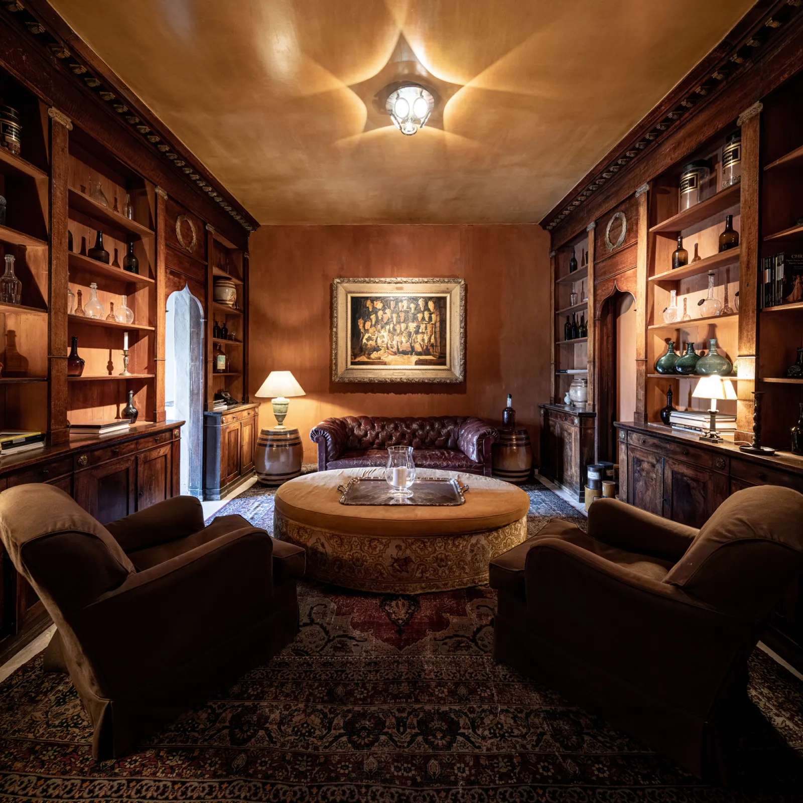 Luxurious library at Château du Tertre in Bordeaux featuring mahogany woodwork, velvet armchairs, central round table, shelving displays, framed portrait, and elegant lighting fixtures.