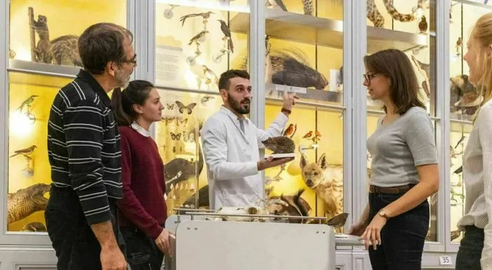 A museum specialist in a white coat explains taxidermied specimens to a group of visitors in front of museum display cases filled with stuffed animals and butterflies.