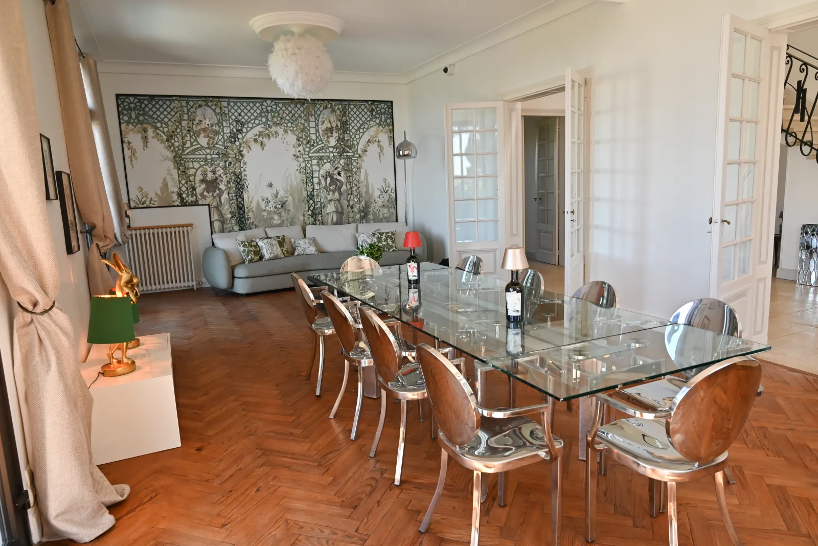 Elegant interior of Chalet des Perchés in Bordeaux: dining area with glass table and wooden chairs, adjacent living room with grey sofa, herringbone parquet, Asian wall decor and contemporary furniture