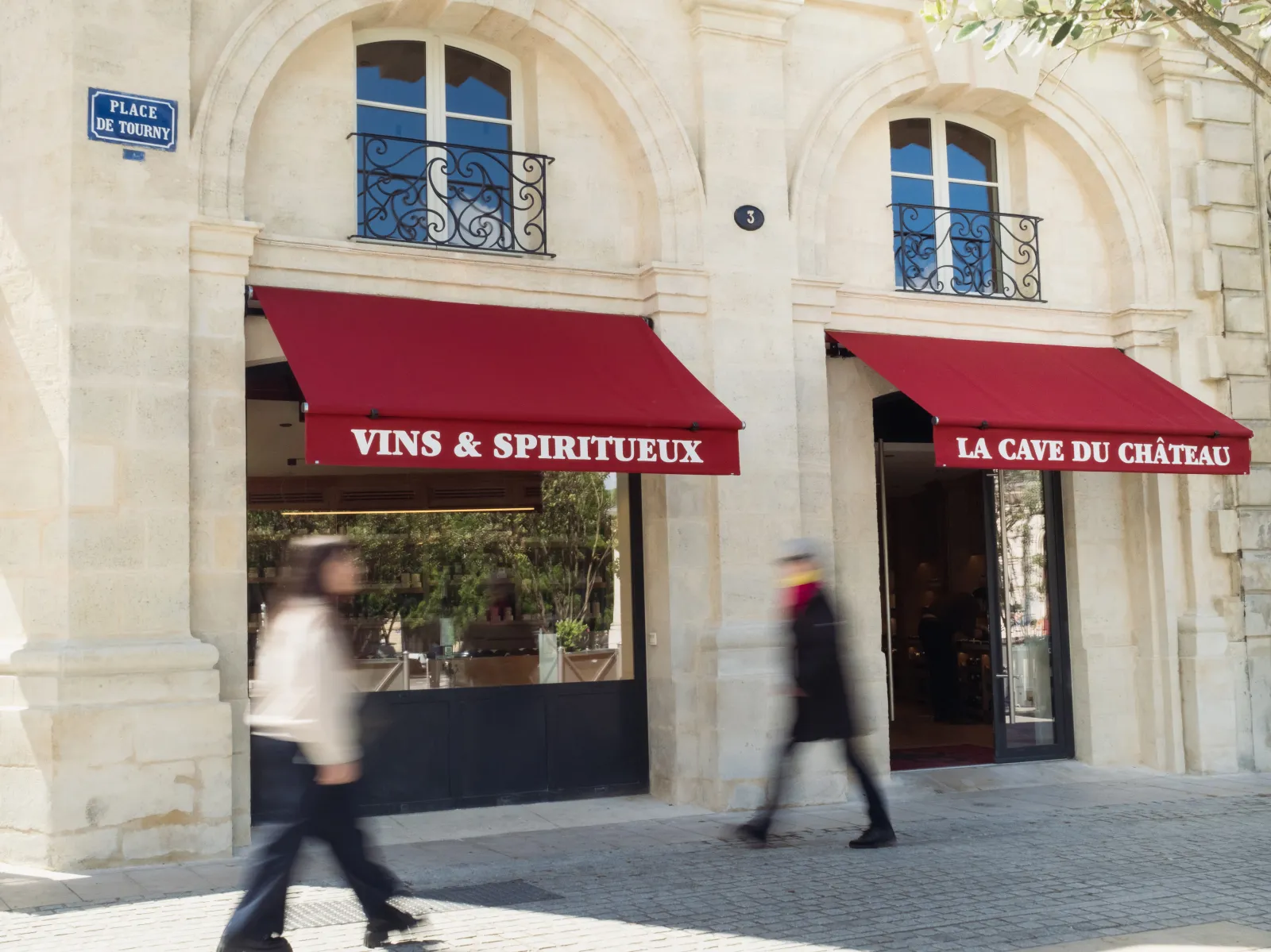 Storefront of a wines and spirits shop 'La Cave du Château' in Bordeaux, located on Place de Tourny, featuring two display windows with red awnings and two blurred pedestrians walking on the paved square.
