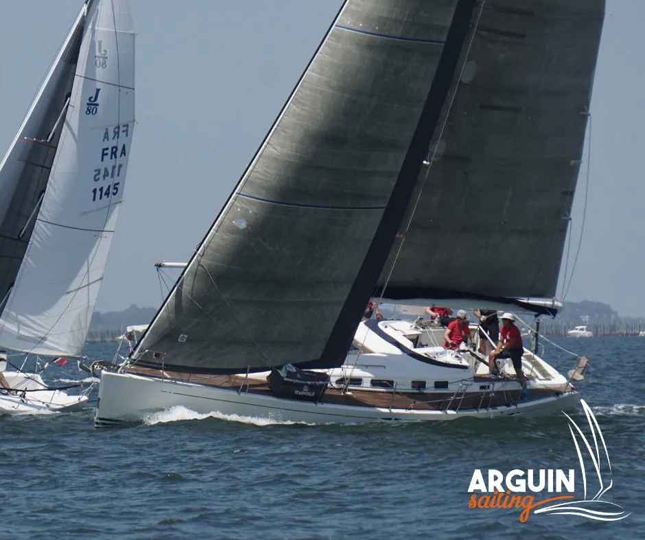 White sailing yacht with black sails cruising on the Gironde estuary near Bordeaux, crew in red onboard with another sailboat visible in the background.