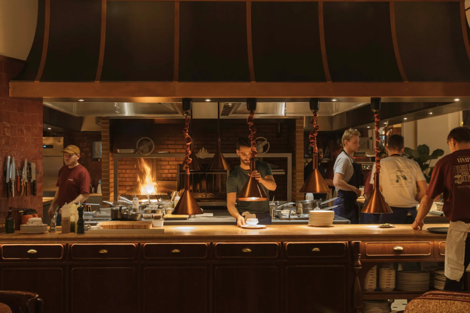 Interior of Brasserie Vinatier in Bordeaux featuring a long wooden counter, chefs cooking in an open kitchen with a roaring fireplace, copper pendant lights, and diners at the bar