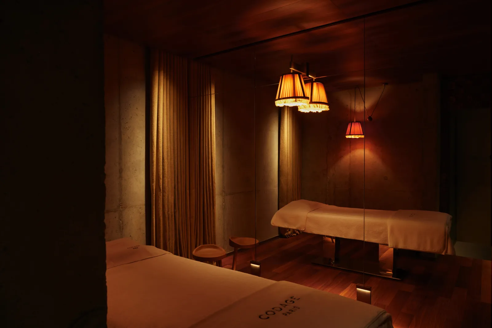 Elegant massage room at Spa Mondrian in Bordeaux featuring two white massage tables, golden ambient lighting, suspended red and gold lamps, beige curtains and wall mirror.