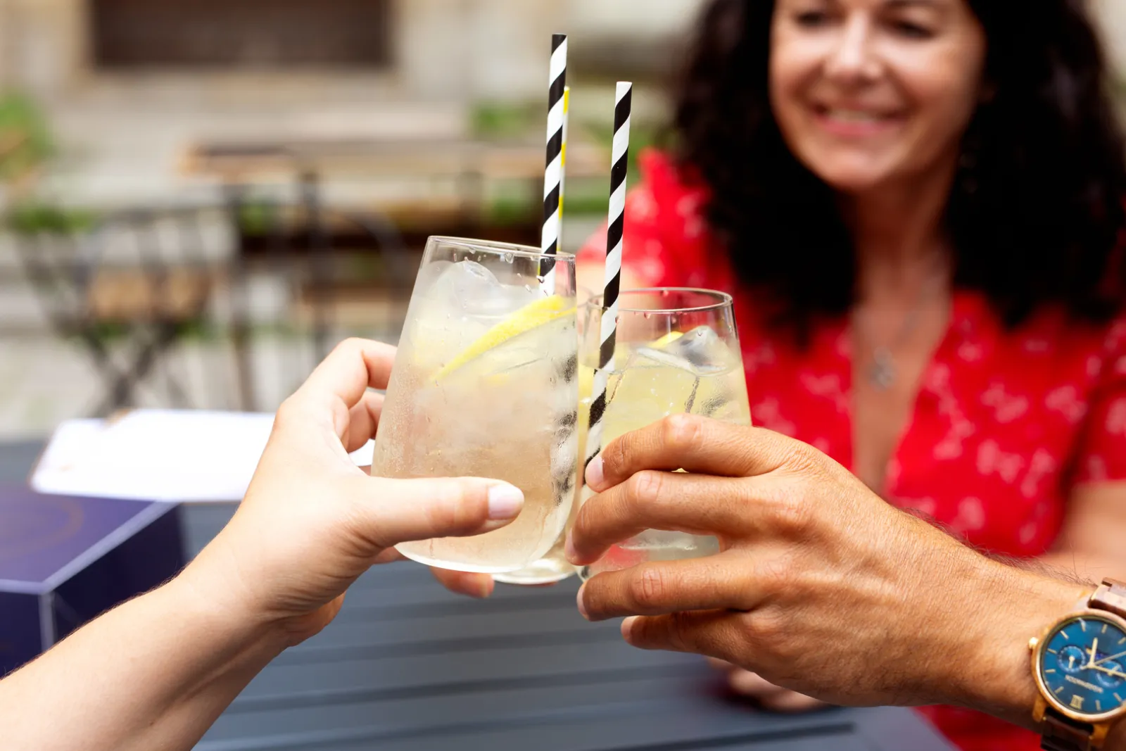 Two glasses of iced lemonade with black and white striped straws held by two smiling people during an outdoor celebration in Bordeaux.