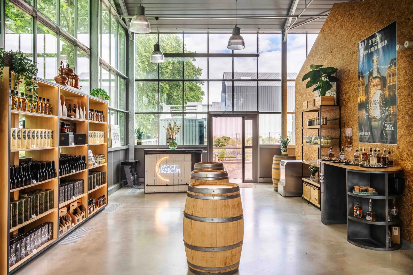 Modern interior of Moon Harbour distillery in Bordeaux featuring wooden shelves with bottles, oak barrels on the floor, black counter, large windows overlooking a garden, and cork walls.