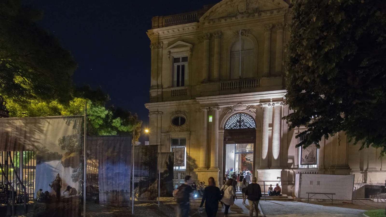 Illuminated façade of the Bordeaux Museum of Fine Arts at night, featuring classical columns and portico, surrounded by visitors and lit trees.