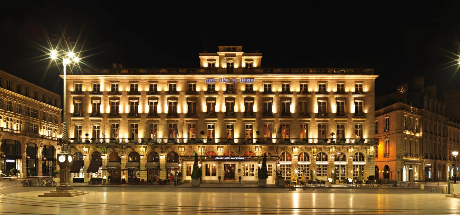 Night view of the Grand Hôtel de Bordeaux with its illuminated neoclassical facade, columns, and ground-floor arcades overlooking an empty paved square.
