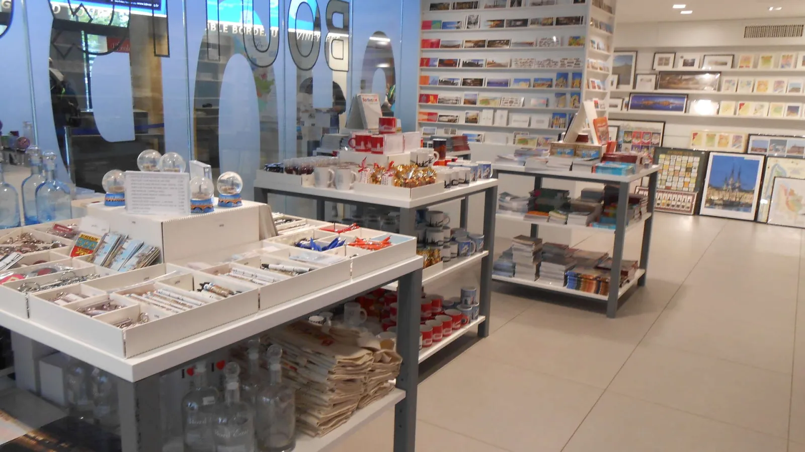 Interior of the Bordeaux Tourism Office shop featuring display counters with souvenirs, books and travel guides, and wall shelves filled with products and tourist documentation.