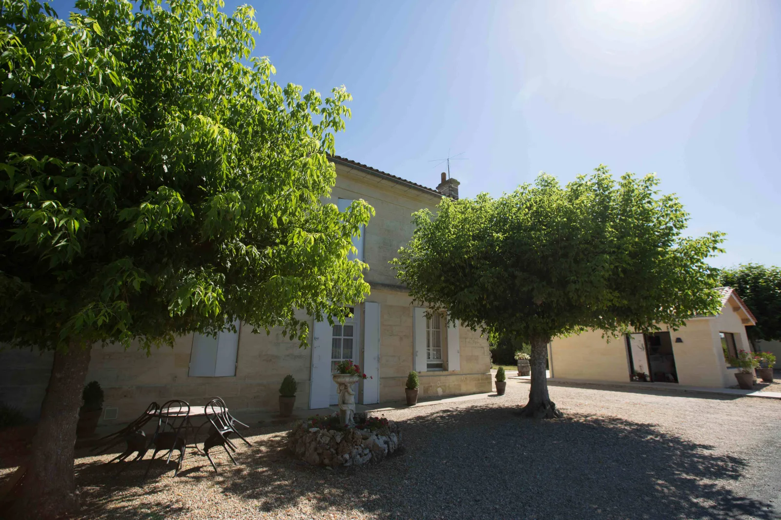 Château Champion in Bordeaux, a stone manor house with white shutters, surrounded by lush green trees and a gravel courtyard, under clear blue sky.