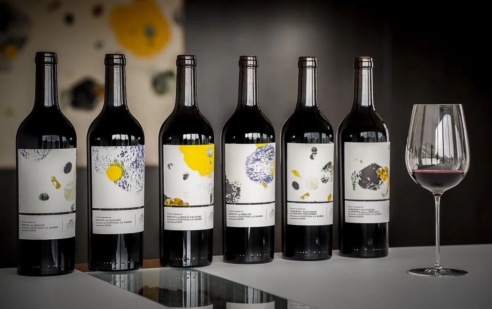 Six bottles of red wine from Château La Garde in Bordeaux lined up with a filled wine glass against a white background with yellow and black paint splatters.