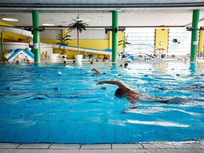 Swimmer performing freestyle stroke in an indoor pool at Pessac Aquatic Stadium in Bordeaux, with other swimmers and pool facilities visible in the background.