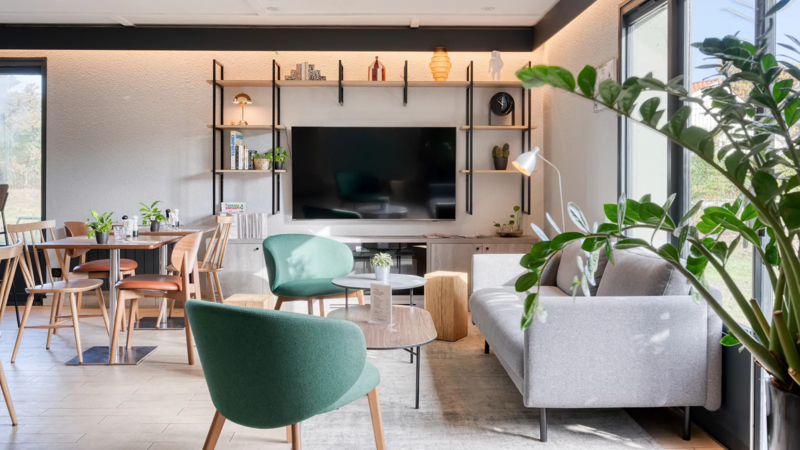Bright modern living room featuring a grey sofa, green armchairs, wall-mounted TV with integrated shelving, potted plants, and wooden dining table at Campanile Nature Bordeaux hotel.