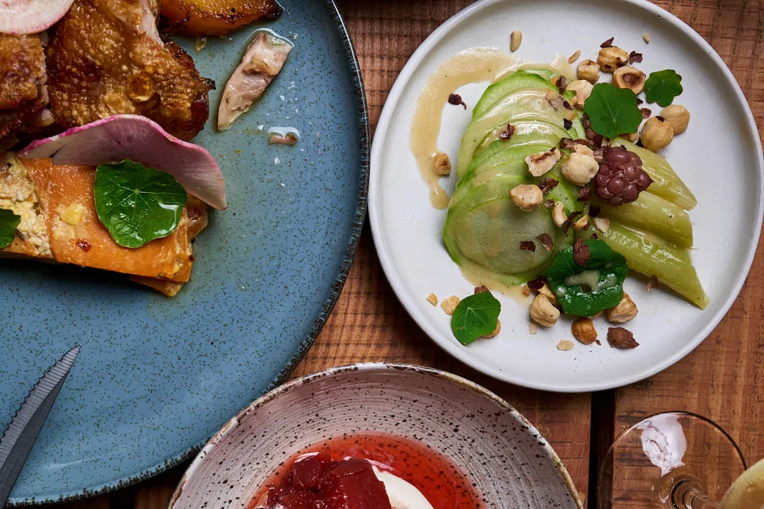 Three gourmet dishes: roasted fish with herbs on blue plate, pear with avocado and nuts on white plate, and red sauce on gray plate.