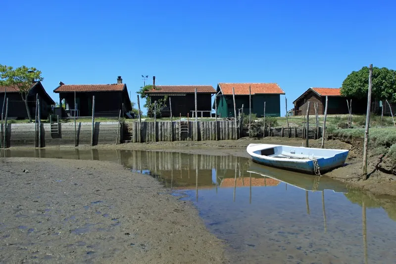 Traditional wooden fishing cabins with red tile roofs along the waterfront on Bird Island near Bordeaux, with a blue and white fishing boat moored in the foreground