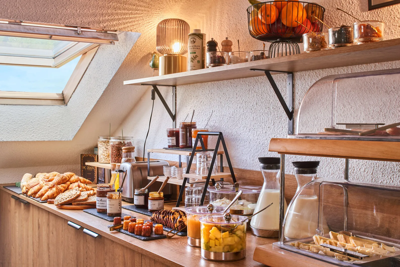 Elegant breakfast buffet featuring pastries, fresh fruits, cereals, jams and orange juice in a bright attic room with wooden shelves and warm décor.