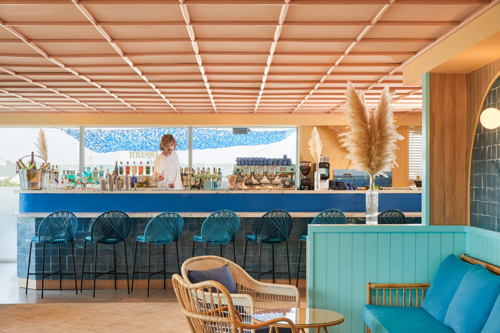 Elegant Tchanqué bar in Bordeaux featuring a blue counter, turquoise bar stools, white slatted ceiling with integrated lighting, pampas grass decoration, and large windows.