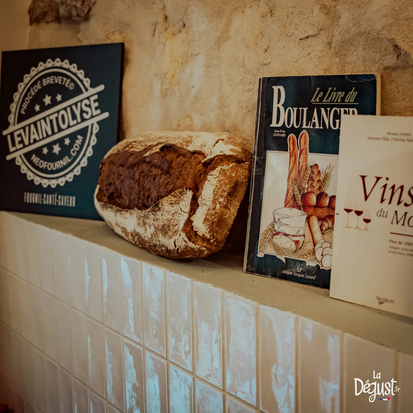 Artisanal French bread displayed on a shelf next to books about bread and Bordeaux wine, with a 'Le Vaintolyse' sign in the background, in a rustic stone-walled setting.