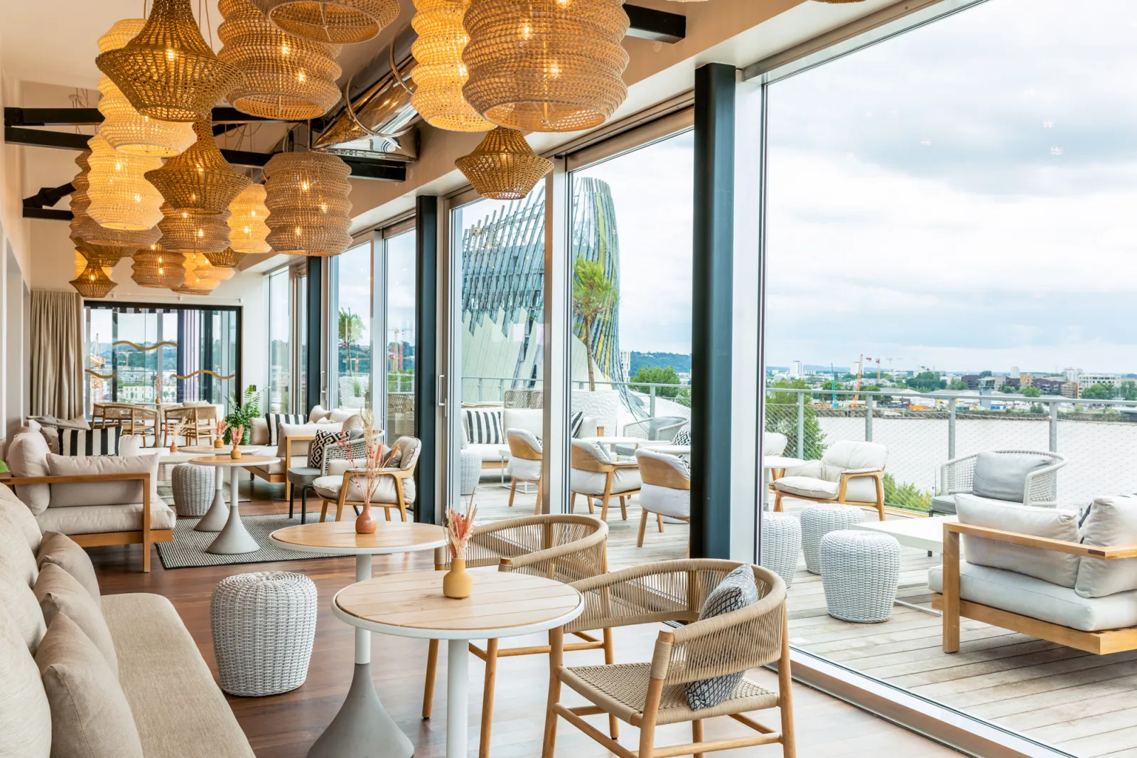 Modern restaurant or bar interior with panoramic views of the Garonne River and Bordeaux, featuring golden chandeliers, wooden and wicker designer furniture, large floor-to-ceiling windows and outdoor terrace.