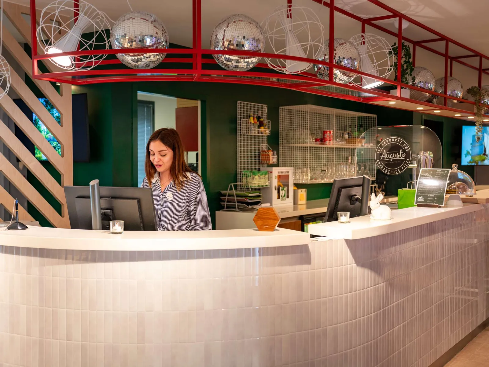 Reception desk of Ibis Styles Bordeaux Centre Gare with staff member, modern decor in green and white tones, silver disco balls hanging under red framework