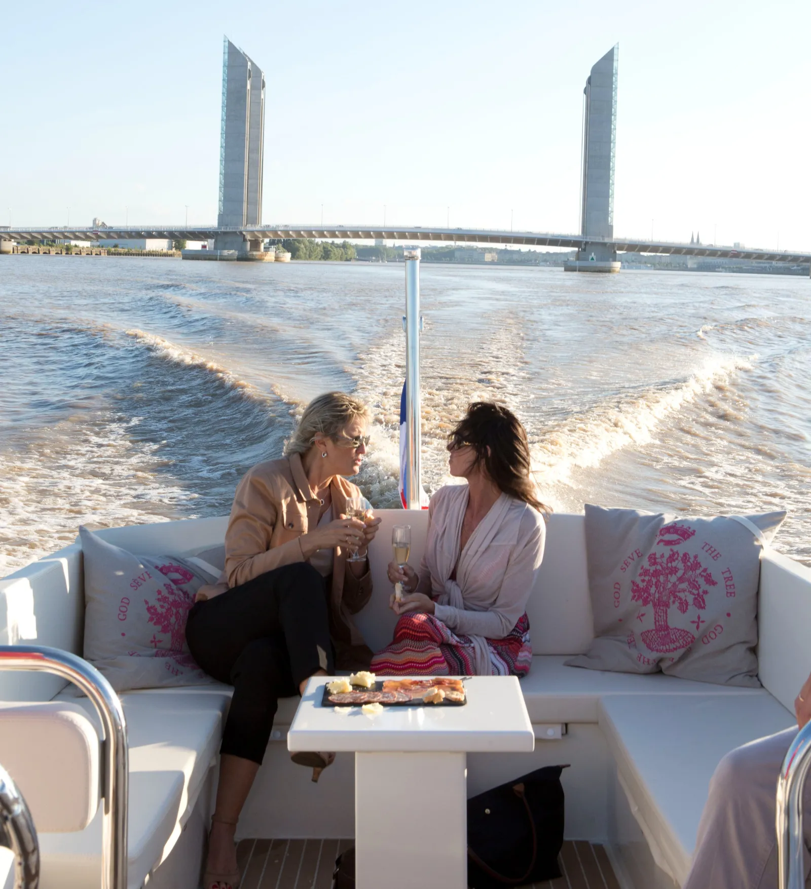 Two women sitting on a white sofa in a water taxi boat in Bordeaux, facing the iconic Stone Bridge with its two towers, the Garonne River in the background on a sunny day.