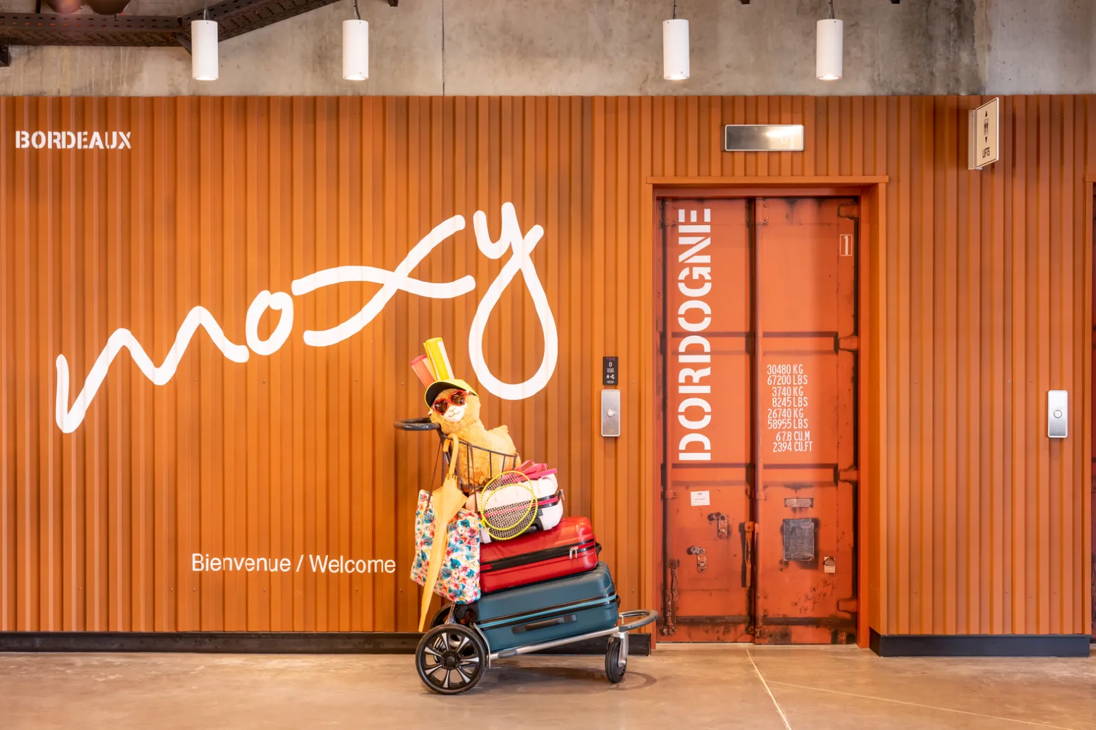 Entrance of Moxy Bordeaux hotel featuring white neon lettering spelling 'Moxy', a colorful illustration of a person with stacked luggage, and 'Bienvenue / Welcome' text on an orange corrugated metal facade.