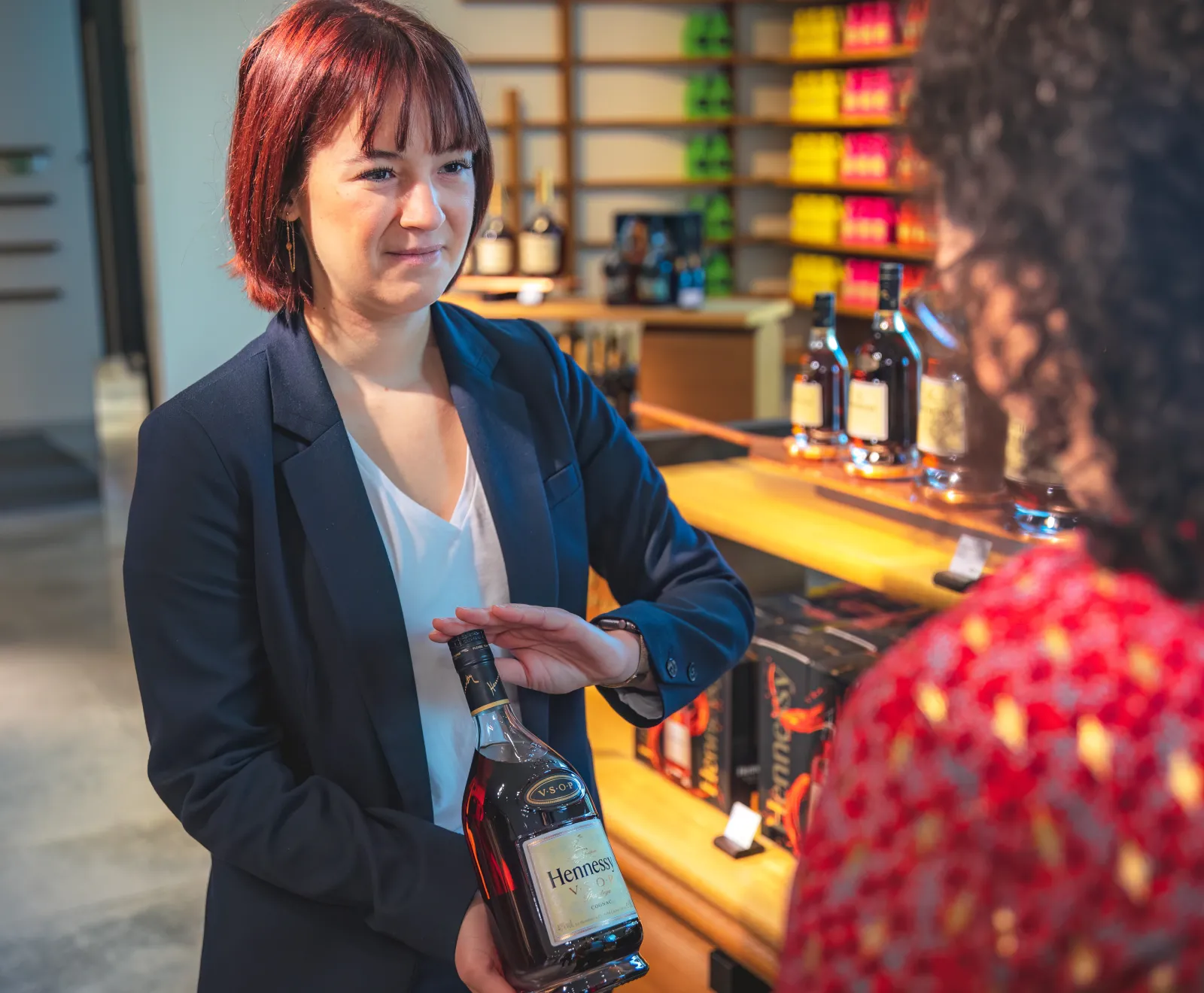 A red-haired woman in a navy blazer holds a Hennessy bottle while presenting in a Bordeaux cognac bar, with colorful shelves and bottles visible in the background.