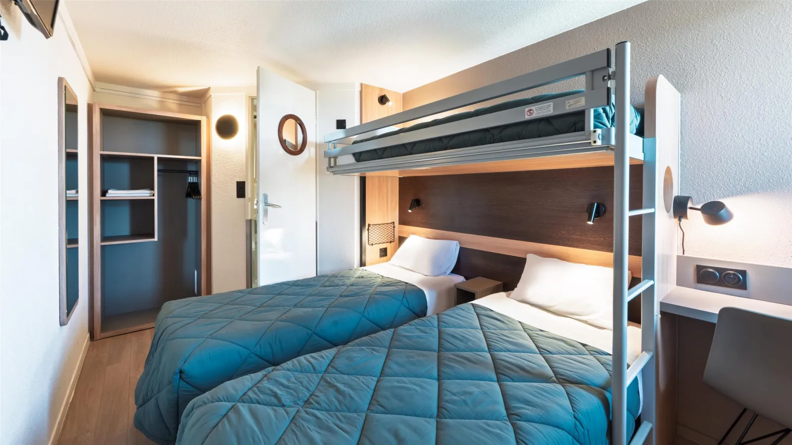 Modern hotel room featuring a gray metal bunk bed with blue bedding, integrated storage, wall-mounted air conditioning, and compact furniture in a minimalist style.
