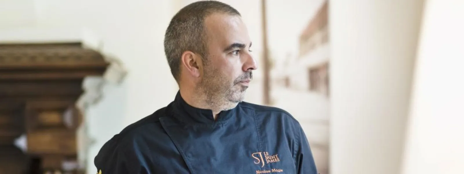 Portrait of a chef wearing a navy blue chef's jacket with the Saint-James Bordeaux logo, looking to the right in a hallway of the restaurant.