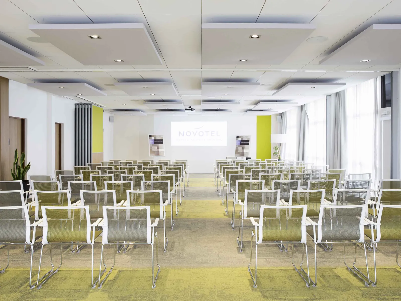 Modern conference room at Novotel Bordeaux Lac featuring rows of white and green chairs facing a projection screen displaying the Novotel logo, with a coffered ceiling with integrated lighting and large windows