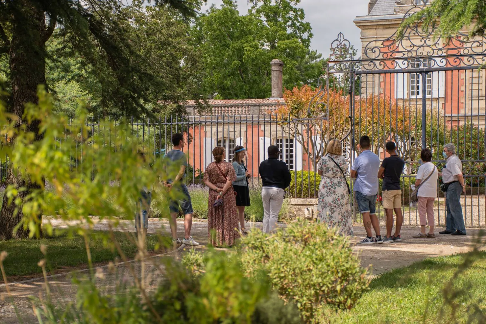Visitors gather in the gardens of Château Saint Ahon in Bordeaux, surrounded by greenery and facing the red brick historic facade with ornate iron gates.