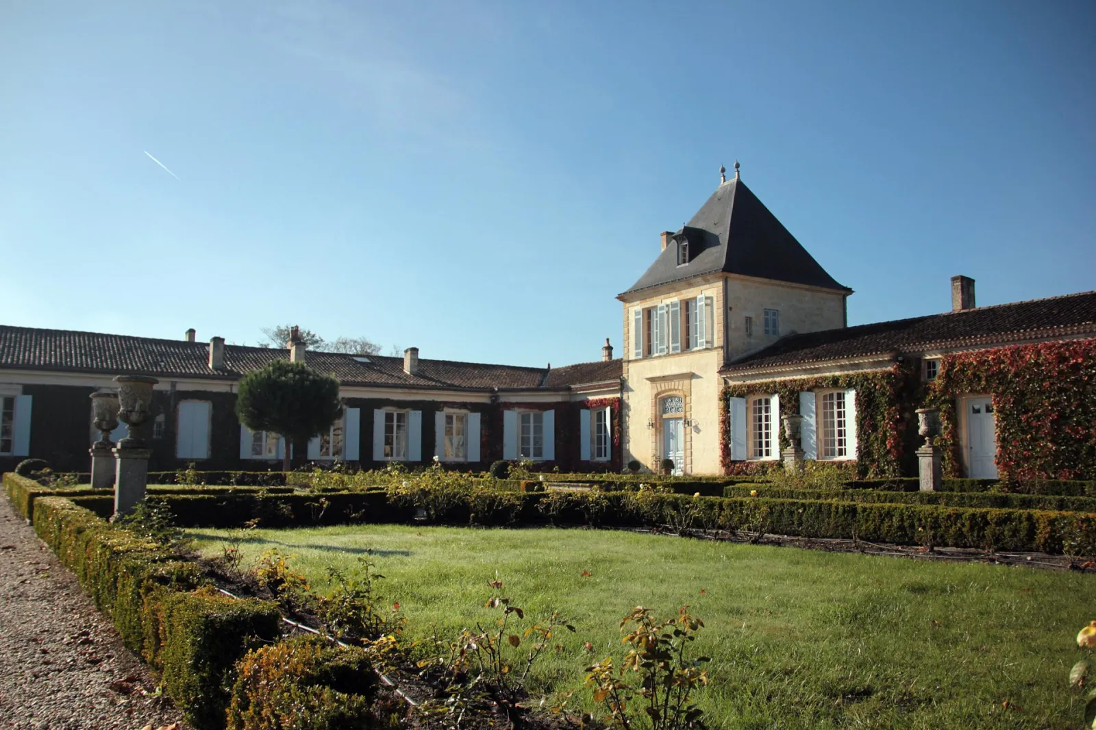 View of Château Paveil de Luze in Bordeaux, featuring a central mansard tower, white stone buildings with blue shutters, surrounded by manicured grounds with lawns and box hedges under clear skies.