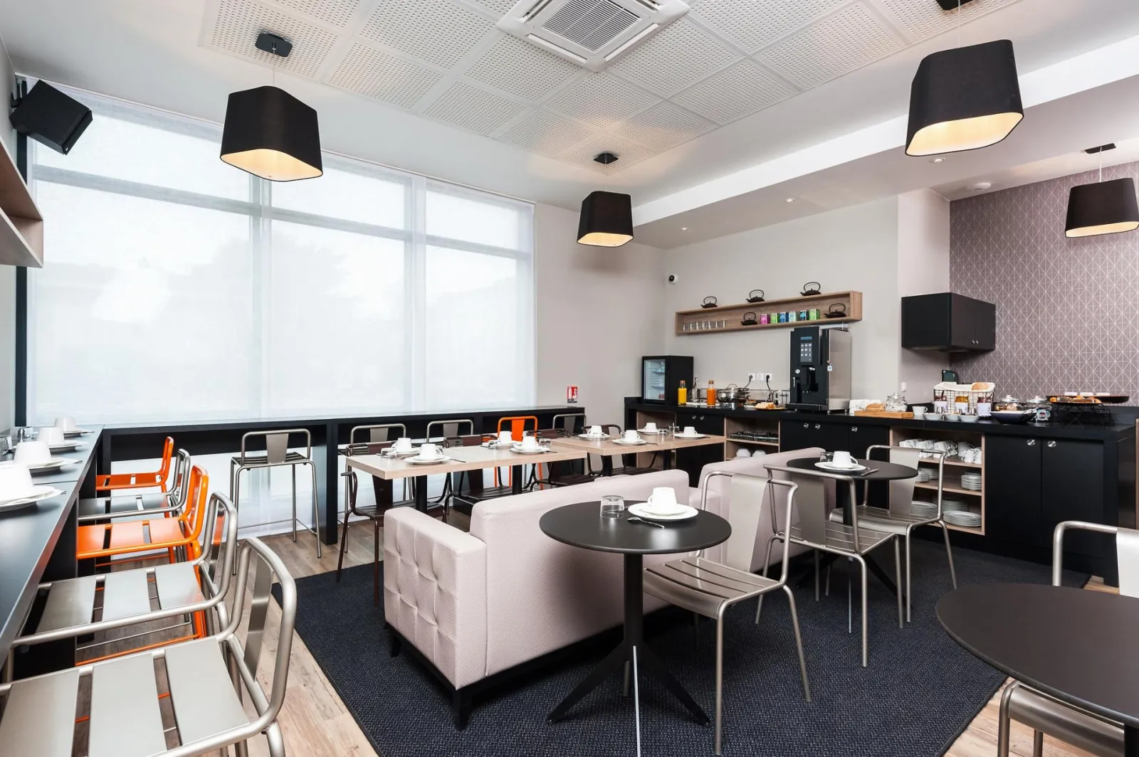 Modern, bright restaurant space featuring black round tables, gray and orange chairs, pale pink sofa, open kitchen with black appliances, large windows, white ceiling with black pendant lights and geometric mauve wallpaper.