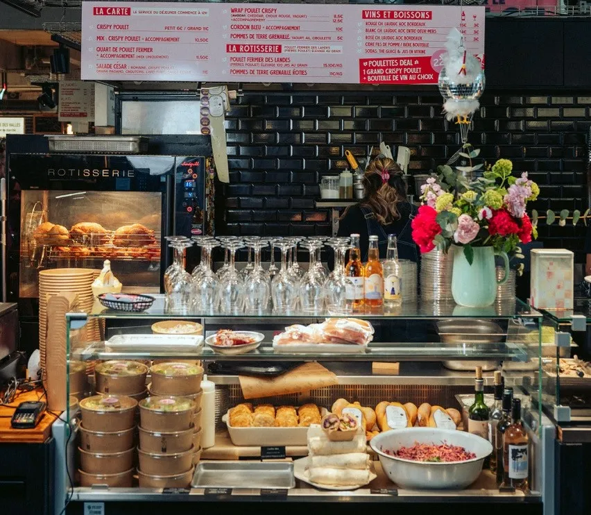 Storefront of Rôtisserie Poulettes in Bordeaux featuring a display case with roasted chickens, cured meats, fresh bread and sides, menu boards above, wine glasses and bottles on the counter, decorative flowers in the foreground.