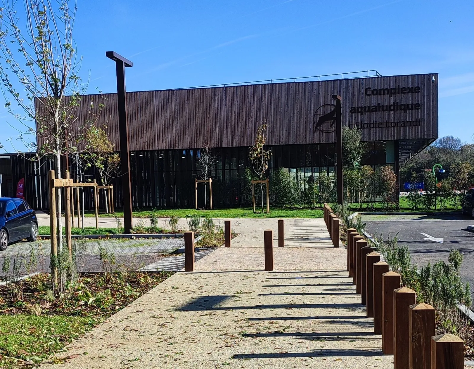 Entrance to the Elodie Lorandi water complex in Bordeaux featuring a modern wooden and steel building, paved pathway with bollards, trees and green spaces