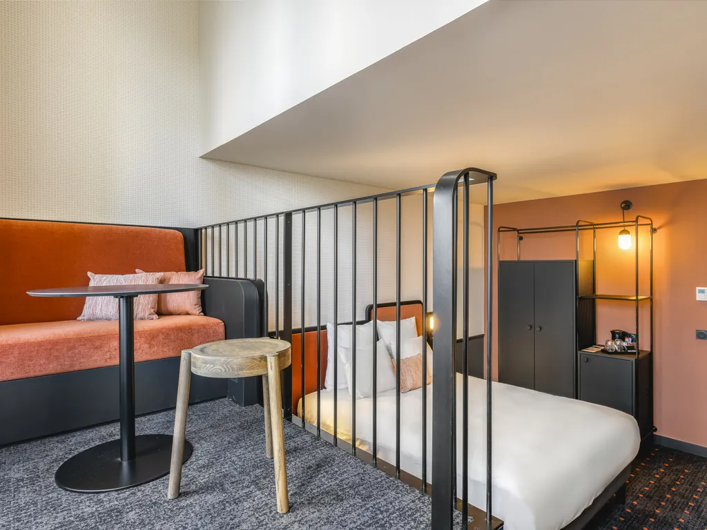 Modern hotel room at Mercure Bordeaux Gare Atlantic featuring a queen-size bed separated by metal railings, orange sofa, black furniture, and contemporary warm-toned design.