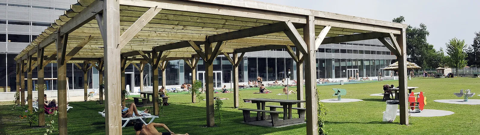 Outdoor relaxation area of the Villenave d'Ornon Olympic pool featuring wooden pergolas, green lawn, picnic tables and visitors relaxing under shaded structures.