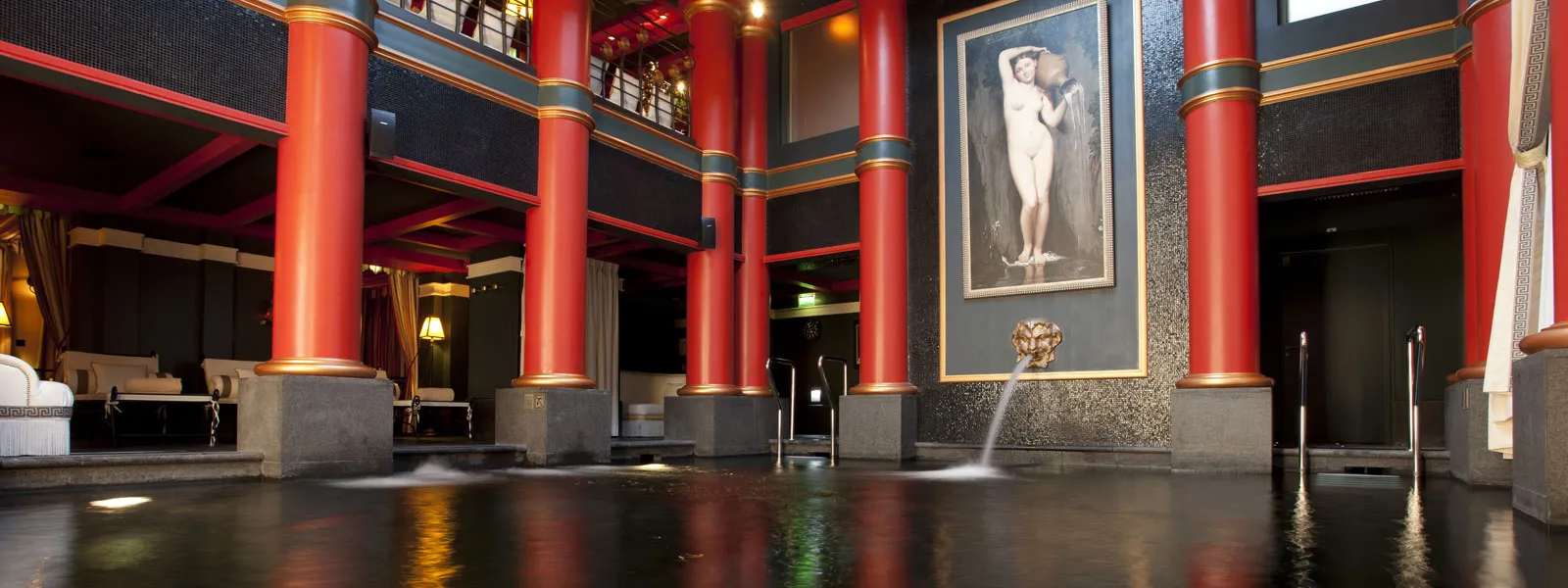 Interior of Guerlain Spa in Bordeaux featuring red ornamental columns, reflective floor, central fountain, and classical framed portrait on textured gray wall.