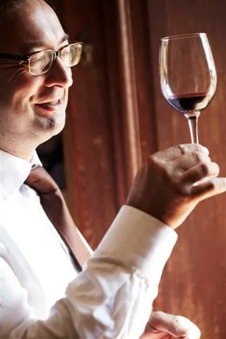 A man wearing glasses and a white shirt carefully examines a glass of red wine against a burgundy curtain background, at Château La Haye in Bordeaux.
