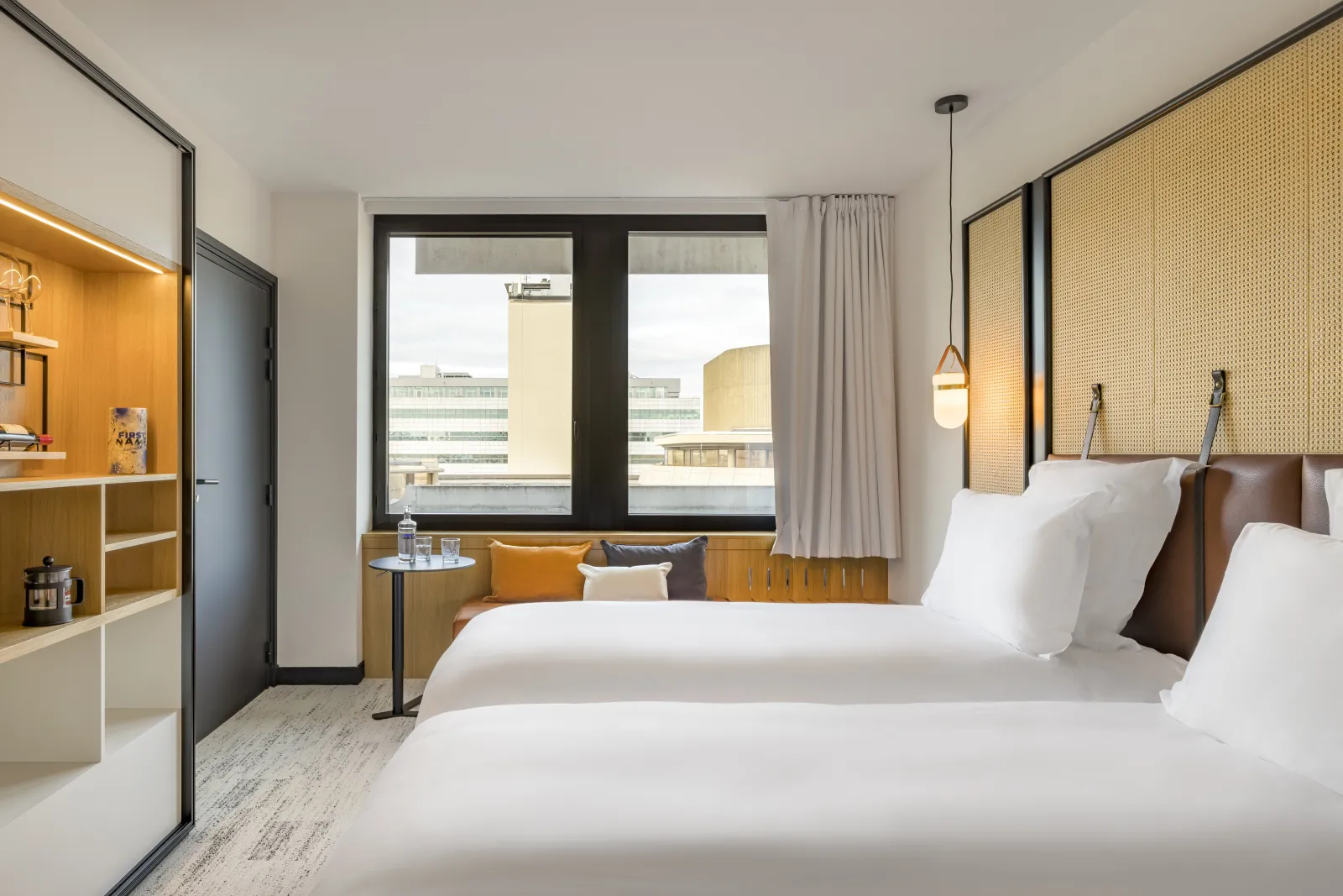 Modern hotel room featuring two queen beds with rattan headboards, gold accents, and contemporary furnishings. Large window overlooking Bordeaux cityscape, with seating area and small table.