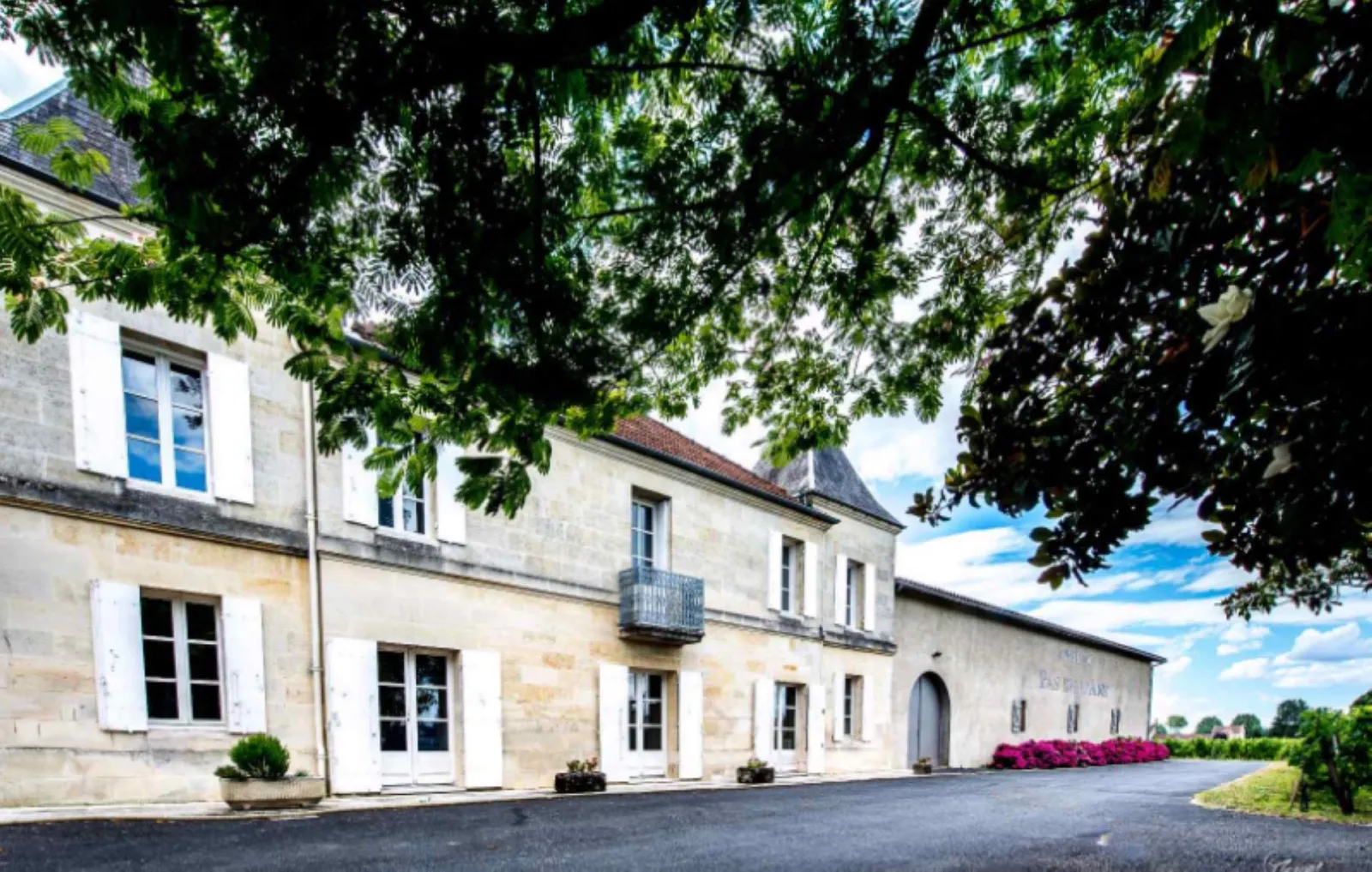 Facade of Château Pas de l'Âne in Bordeaux, white stone building with white shutters, blue windows, wrought iron balcony, paved courtyard shaded by trees
