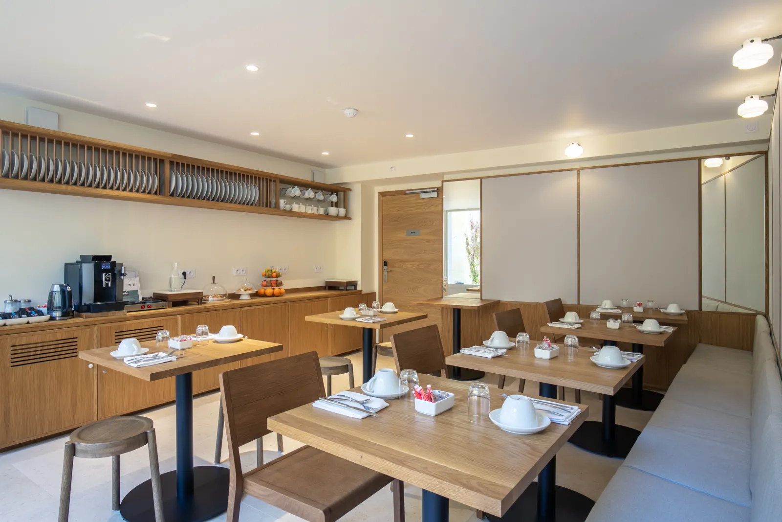 Elegant breakfast room at Badon Boutique Hotel in Bordeaux, featuring wooden tables, brown chairs, white tableware, light wood kitchenette and open shelving.