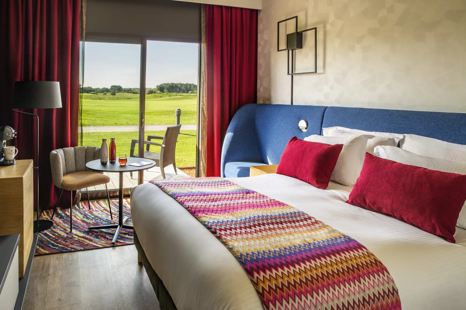 Modern hotel bedroom with king-size bed with blue headboard overlooking green fields through large glass doors, red curtains, red pillows and colorful patterned throw blanket