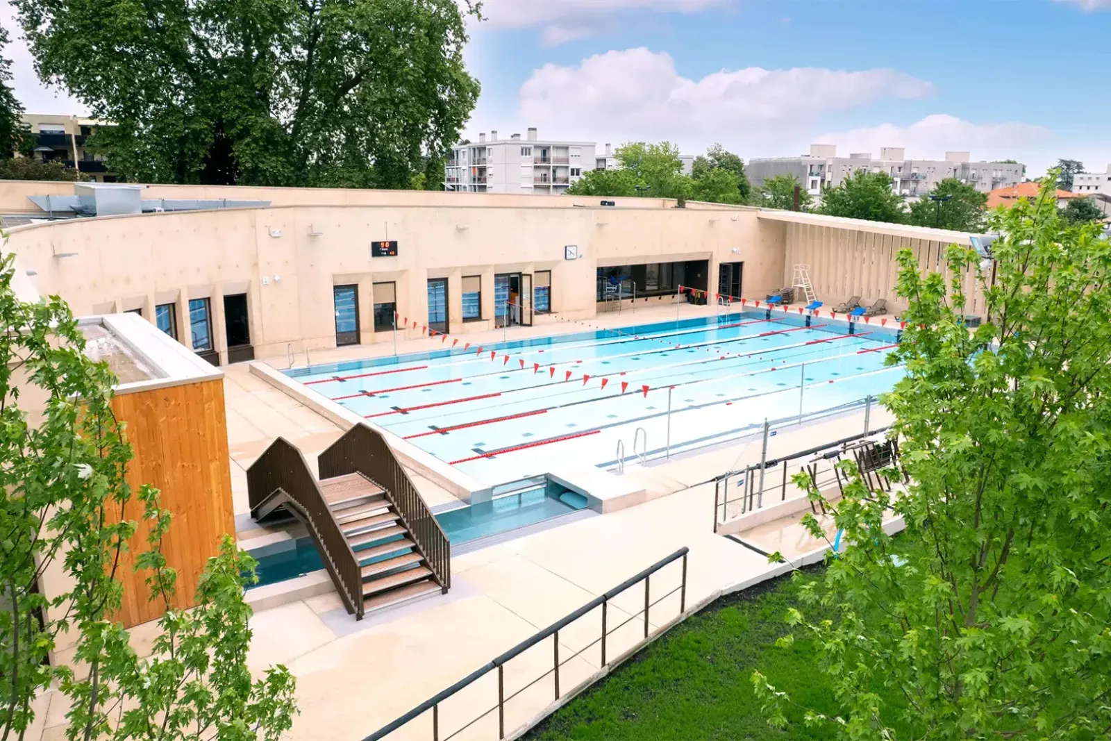 Aerial view of Jean Zay Swimming Pool in Bordeaux featuring an Olympic-size pool with blue and red lanes, surrounded by a modern beige building and green trees