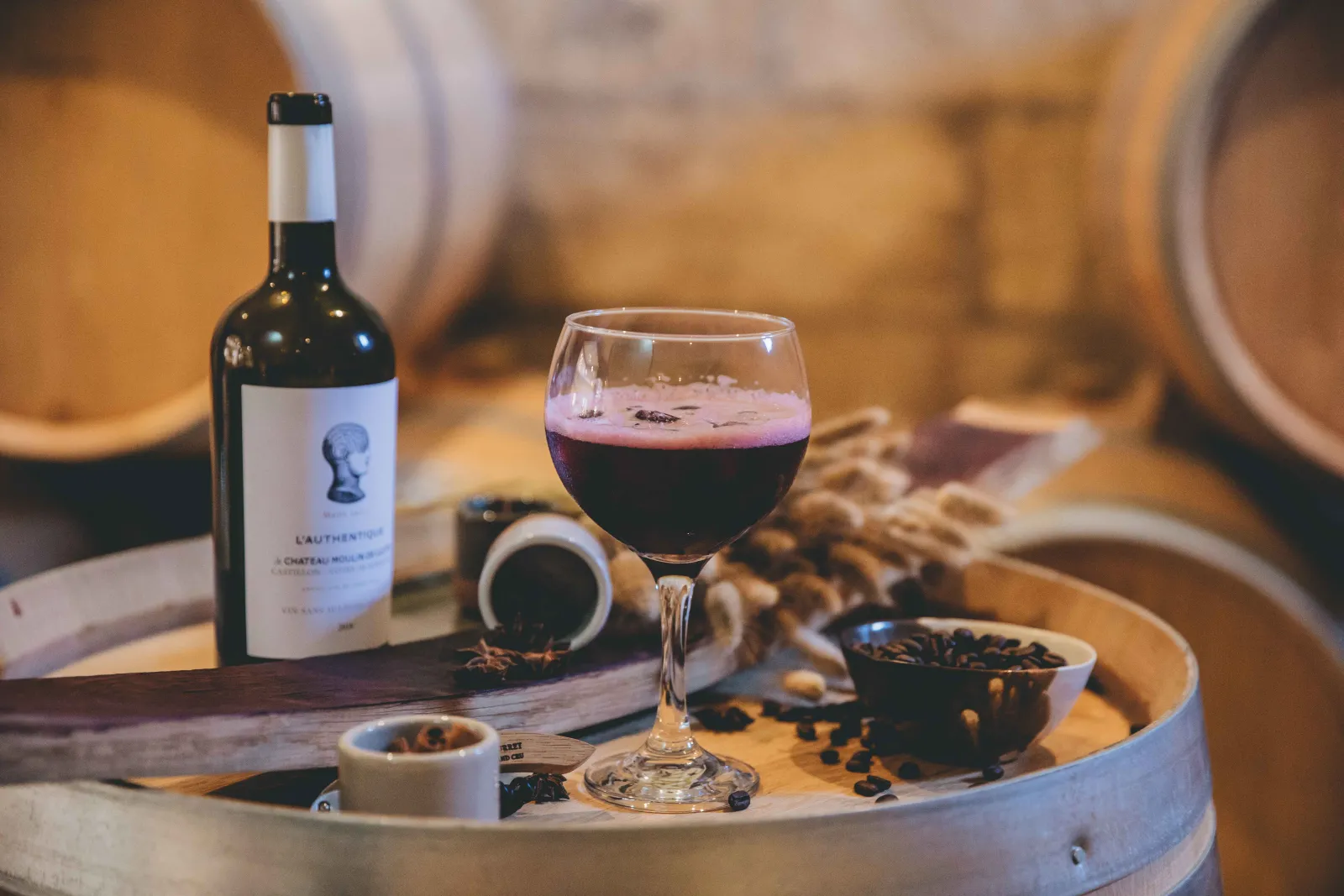 Glass of red Bordeaux wine from Château Ambe Tour Pourret with bottle, grapes, and oak barrels visible in the cellar background.