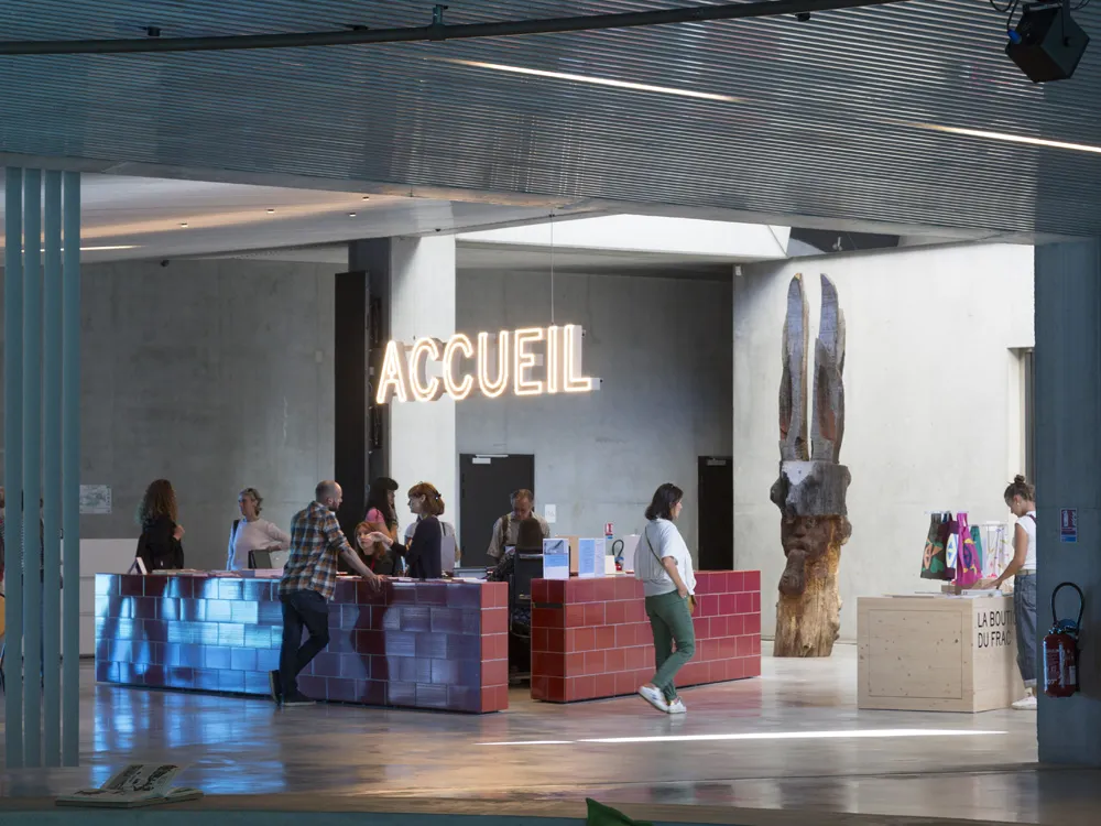 Reception area of FRAC Nouvelle-Aquitaine MECA in Bordeaux featuring a blue and red brick reception desk, wooden sculpture and visitors in a bright contemporary space