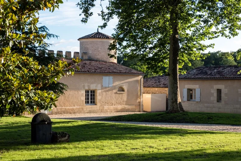 View of Château Lafaurie Peyraguey in Bordeaux featuring its distinctive round tower, ochre stone buildings, surrounded by lush greenery and ancient trees